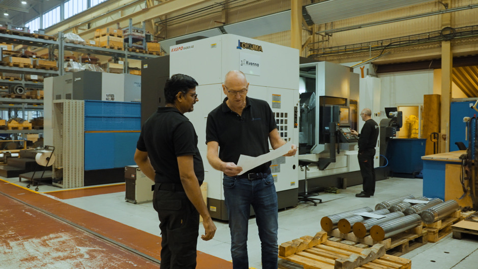 To menn i svart arbeidstøy står i en industrihall og ser på et dokument. I bakgrunnen er en stor maskin med teksten ‘Okuma’, samt hyller fylt med utstyr og en medarbeider som jobber ved en maskin. Flere metallstenger ligger stablet på paller i forgrunnen.