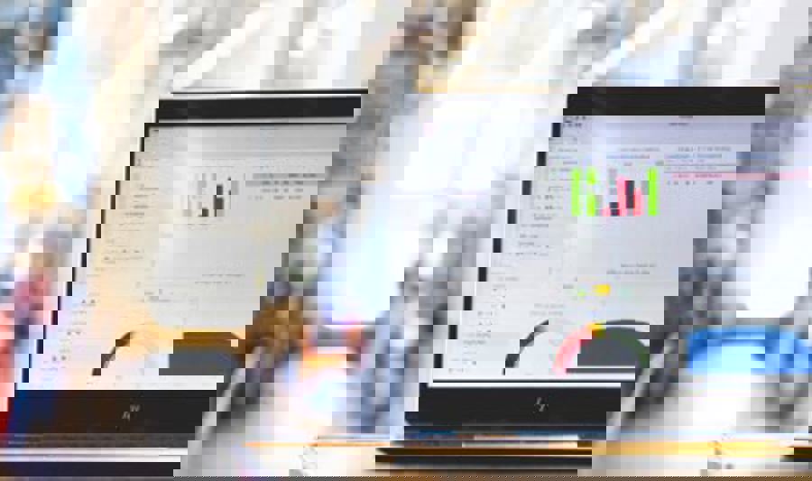 En bærbar PC med et dashbord som viser grafer og dataanalyse står på et bord i et industrielt lager- eller produksjonsmiljø. Fargerike diagrammer og grafer vises på skjermen, mens bakgrunnen er uskarp med reoler og utstyr.