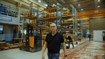 Man walking through an industrial warehouse with shelves filled with equipment and tools, a forklift in the foreground, and workers in the background.